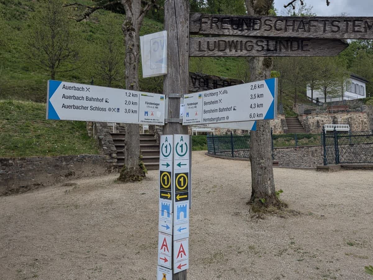 Ein detaillierter hölzerner Wanderwegweiser mit zahlreichen blauen und weißen Schildern, die Richtungen und Entfernungen (Auerbach, Bensheim, Kirchberg etc.) angeben, sowie Wanderzeichen (A1, Burgensteig), davor eine Steintreppe.